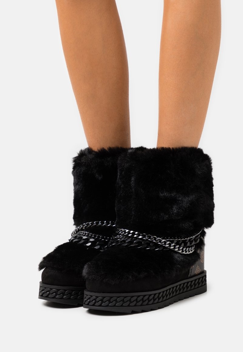 Bottines en fourrure noire avec des semelles épaisses, des chaînes décoratives enroulées autour et des talons à motifs portées sur des jambes nues.