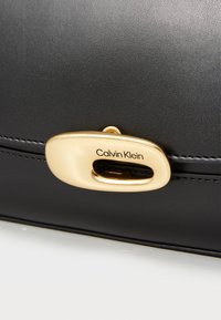 Černá kožená taška s zlatou oválnou sponou s vyrytým nápisem "Calvin Klein", hladkým povrchem a jemným detailem prošívání.