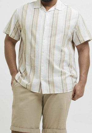 Mann trägt ein kurzärmliges beige-weiß gestreiftes Button-up-Hemd mit beigen Shorts, steht mit den Händen in den Taschen.