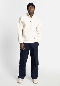 Romige fleecetrekker met een halve rits, kangoeroezak; gecombineerd met marineblauwe cargobroek met meerdere zakken; beige sneakers.