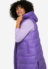 Gilet en mousse violet avec capuche, présentant un design matelassé, des poches latérales et une texture lisse, porté par-dessus un haut à manches longues lavande.