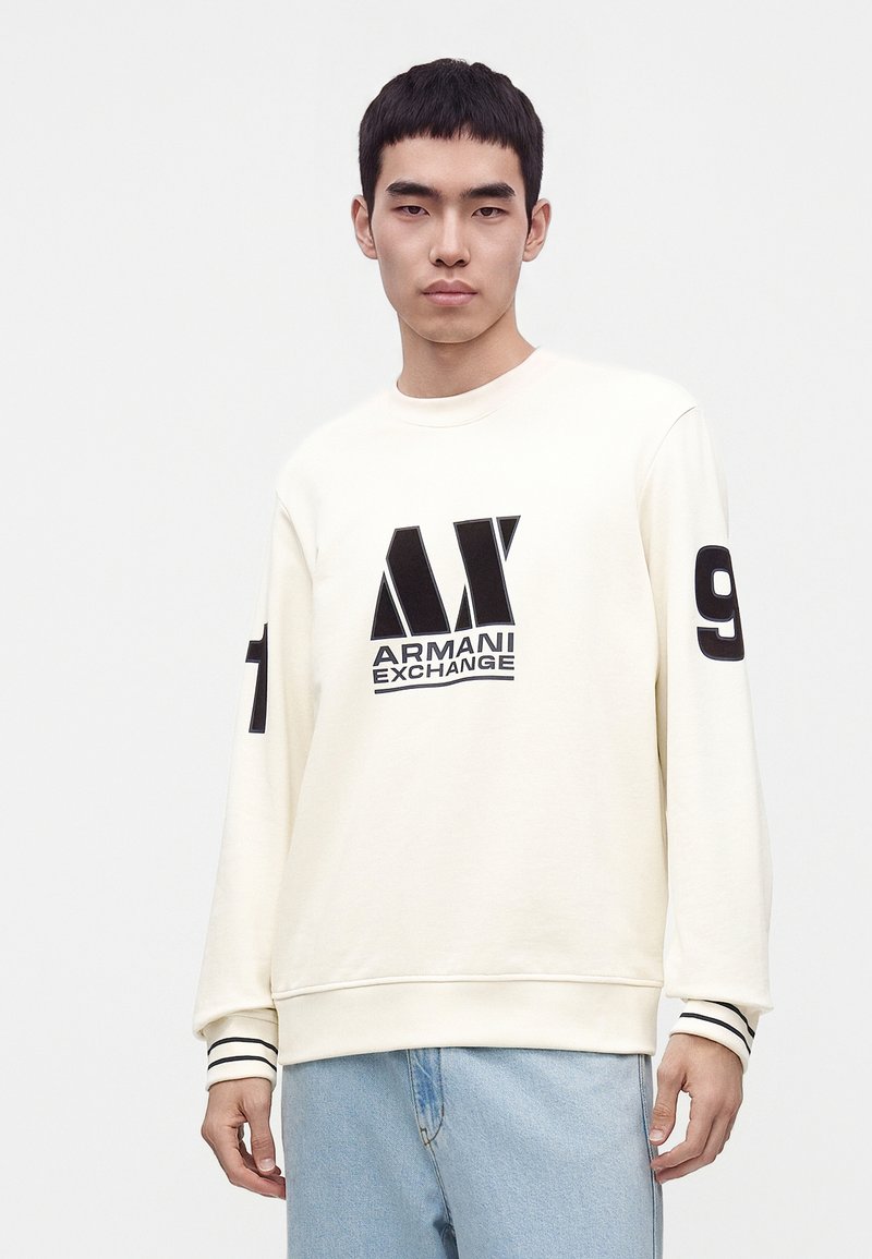 Jeune homme portant un sweat-shirt crème Armani Exchange avec logo noir sur le devant et chiffres sur les manches, associé à un jean bleu clair.