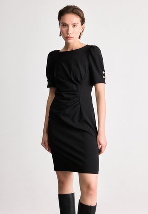 Robe noire avec des manches bouffantes courtes, texture drapée et détails froncés à la taille. Comprend des accents de boutons dorés sur les manches.
