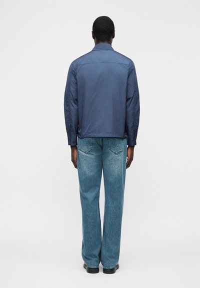 Homme portant une veste bleu marine et un jean droit bleu clair, debout de dos sur un fond blanc uni.