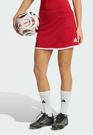 Person in rotem Sportrock hält einen Fußball, trägt weiße Adidas-Socken und schwarze Fußballschuhe mit neongrünen Akzenten.