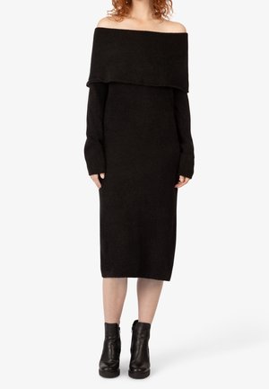 Mujer que lleva un vestido de punto midi negro con hombros descubiertos, mangas largas y botas negras hasta el tobillo, de pie frente a un fondo liso.