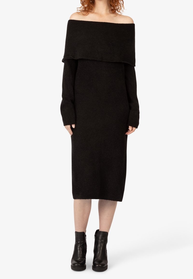 Femme portant une robe-pull midi noire à épaules dénudées avec manches longues et bottines noires, debout devant un fond uni.