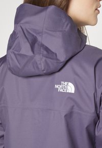 Chaqueta impermeable morada con capucha, con un logo blanco en la parte trasera. Se aprecia una textura lisa y detalles de costuras.