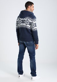 Strikket marineblå hettegenser med hvite geometriske mønstre på øvre bryst, med teksturert stoff og ribbestrikket kant. Båret sammen med mørke jeans og hvite joggesko.