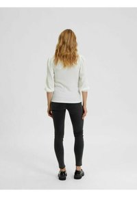 Blusa bianca strutturata con maniche a sbuffo, indossata con jeans neri aderenti e scarpe nere. La vista da dietro mostra la lunghezza e la vestibilità della camicia.