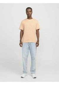 Camiseta beige de manga corta combinada con jeans celestes, de pie sobre un fondo neutro. Zapatillas blancas completan el conjunto.