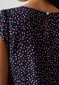 Blouse bleu marine avec un motif dispersé de points blancs et roses, manches plissées et détail dos en forme de goutte fermé par un bouton.