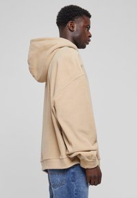 Beige hoodie met een grote capuchon, een losse pasvorm en verlaagde schoudernaden. Zacht materiaal en ribbed cuffs. Draag het met blauwe denim jeans.
