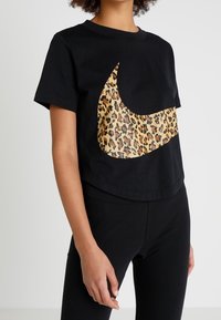T-shirt nera a maniche corte in cotone, con il logo Nike a stampa leopardo sul davanti. Indossata con leggings neri.