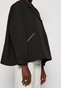 Musta ylisuuri takki, jossa on kaulus, kaksoispainikeet, sivussa vetoketjullinen tasku ja teksturoitu kangas. Päällä vaaleat housut.