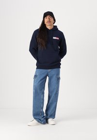 Marineblå hettegenser med frontlogo, kombinert med løse blå denimbukser med sidelommer og hvite sneakers.