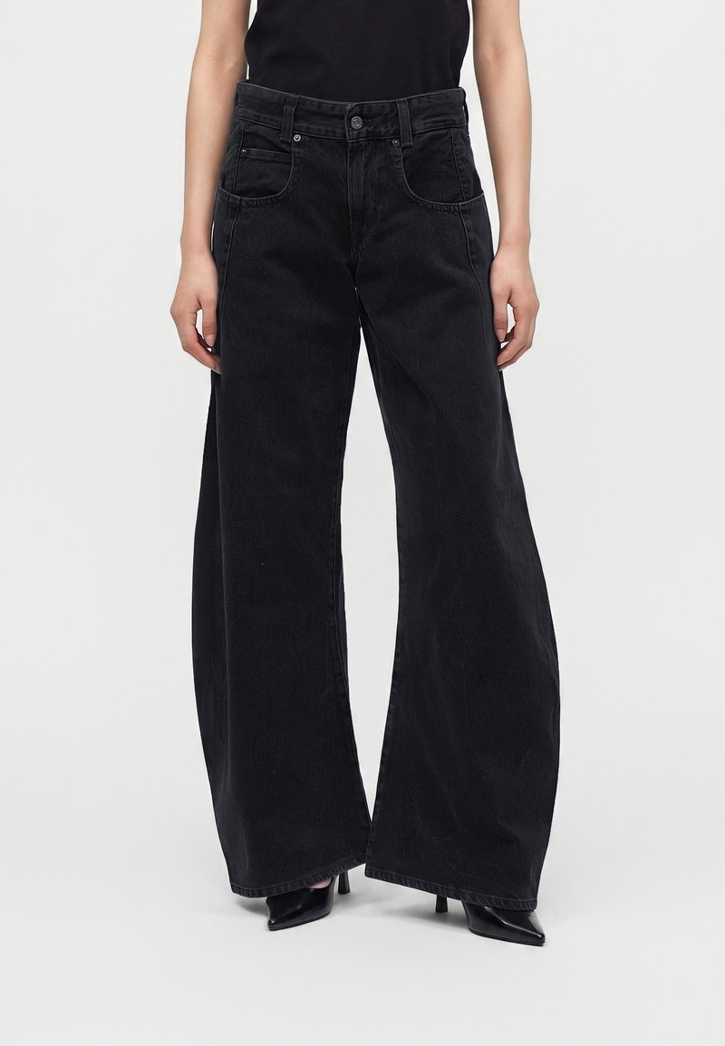 Personne portant un jean noir taille haute à jambes larges et des escarpins noirs à bout pointu, debout devant un fond clair uni.