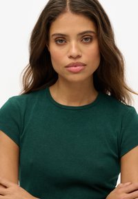 Grøn bomulds-t-shirt med korte ærmer og rund hals. Stoffet er glat med en let stræk. Modellen står med armene over kors, vendt fremad.