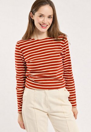 Femme souriante aux longs cheveux bruns portant un haut à manches longues rayé rouge et blanc et un pantalon crème taille haute sur un fond uni.