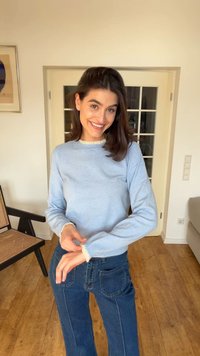 Γυναίκα που χαμογελάει μέσα σε εσωτερικό χώρο, φοράει ανοιχτό μπλε πουλόβερ με λευκή δαντέλα στο τελείωμα και ψηλόμεσα σκούρα μπλε τζιν, όρθια σε ξύλινο πάτωμα.