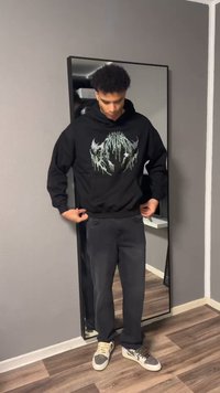 Jeune homme ajuste un sweat à capuche graphique noir, debout devant un grand miroir dans une pièce grise avec un sol en bois.