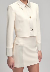 Veste courte couleur crème avec un col pointu, des boutons dorés, et une jupe taille haute assortie avec des poches latérales et une texture lisse.