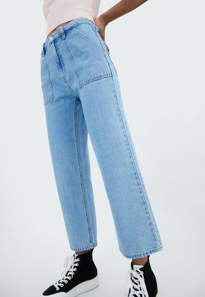 Persoon in lichtblauwe, hoge taille, wijde jeans met grote voorzakken en zwarte high-top sneakers met witte veters.