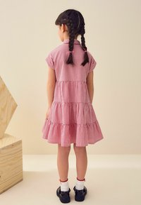 Punainen ja valkoinen ruudullinen mekko lyhyillä hihoilla ja kerrostetulla muotoilulla. Yhdistetty mustiin kenkiin ja valkoisiin sukkiin. Letitetyt hiukset näkyvät takaa.