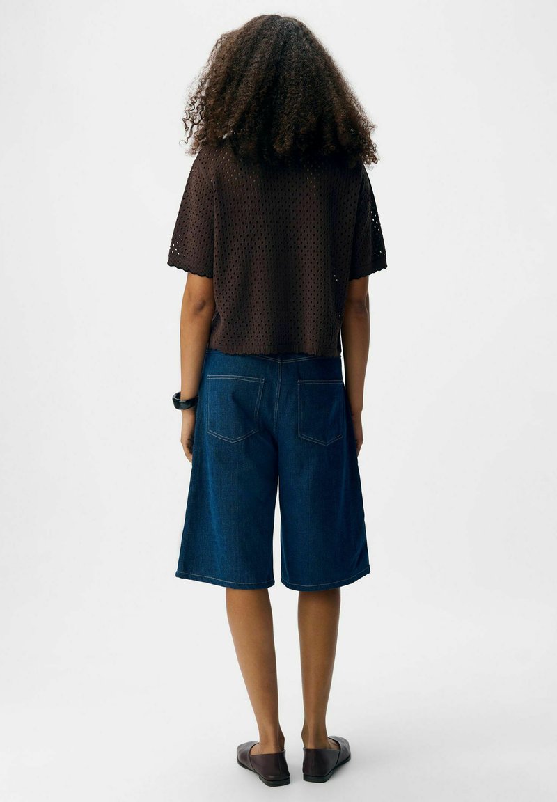 Osoba z kręconymi włosami, ubrana w ciemnobrązową, perforowaną bluzkę z krótkim rękawem, niebieskie szorty dżinsowe do kolan oraz ciemne buty na slip-on.