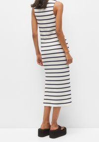 Femme se tenant debout portant une robe midi sans manches blanche avec des rayures horizontales noires et des sandales compensées noires sur un fond blanc uni.