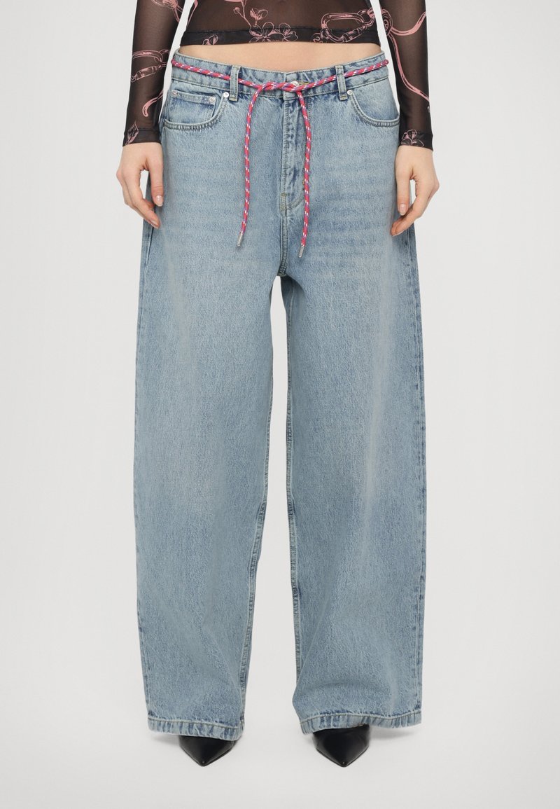 Helle blaue weite Jeans mit hoher Taille, ausgestattet mit einer rot-weißen geflochtenen Kordel und kontrastierenden schwarzen spitzen Schuhen.