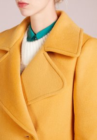 Cappotto di lana giallo con rever a taglio, caratterizzato da una finitura testurizzata. Sotto, è visibile un colletto verde di un maglione lavorato a maglia bianco.