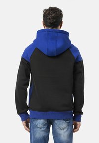 Sweat à capuche avec un corps noir, une capuche bleue et des manches bleues ; doté de poignets et d'un ourlet côtelés, fabriqué en tissu doux et texturé.