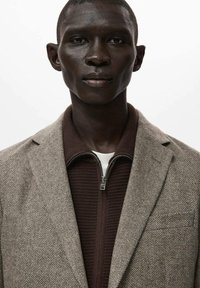 Hnědý blazer s rybí kostí přes tmavě hnědý zipový top. Hladká textura, střih na míru, jedna náprsní kapsa na blazru.