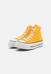 Zapatillas altas de lona amarilla brillante, con cordones blancos y suela de goma. Presentan una franja negra y una puntera texturizada.