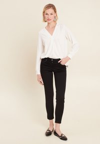 Weiße Longsleeve-Bluse mit V-Ausschnitt und strukturiertem Stoff, kombiniert mit eng anliegenden schwarzen Jeans und schwarzen Ballerinas mit silbernen Hardware-Akzenten.