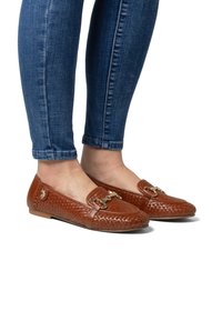 Hnědé pletené mokasíny s plochou podrážkou, se zlatou sponou a malým logem na boku. Kombinováno s modrými úzkými džíny.