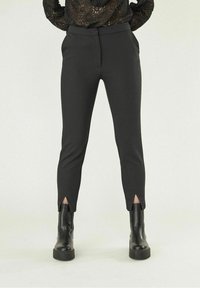 Femme portant un pantalon noir ajusté avec des fentes à l'avant de la cheville, associé à des bottines à talons épais noires et un haut en dentelle à manches longues.