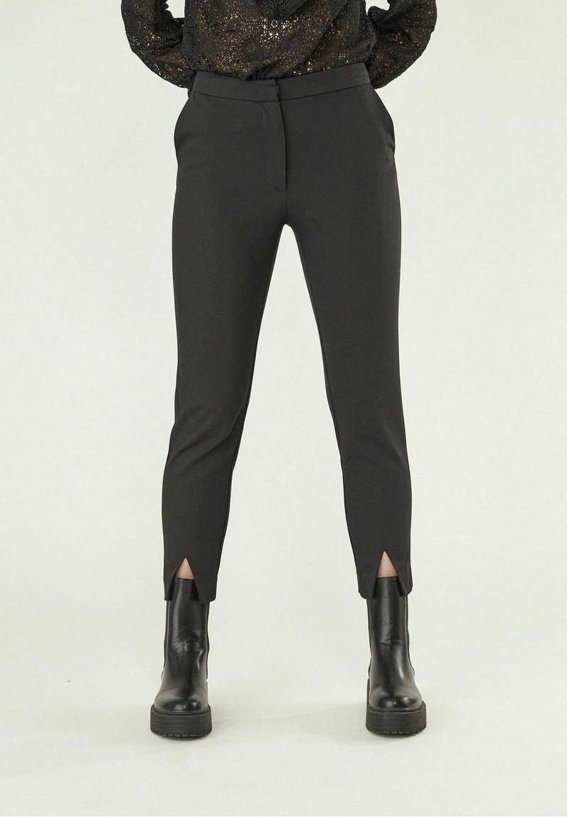 Femme portant un pantalon noir ajusté avec des fentes à l'avant de la cheville, associé à des bottines à talons épais noires et un haut en dentelle à manches longues.