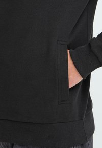 Mano parcialmente en el bolsillo delantero de una sudadera negra con puños y bajo acanalados, contra un fondo claro.