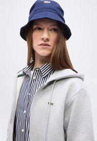 Νεαρή γυναίκα με μακριά μαλλιά, φοράει σκούφο τύπου bucket σκούρου μπλε, ριγέ πουκάμισο με κουμπιά και ανοιχτό γκρι φούτερ με φερμουάρ και μικρό πράσινο λογότυπο.