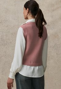 Gilet en tricot rose avec un motif texturé, un ourlet côtelé et des boutons aux épaules, porté sur une blouse blanche, associé à un jean foncé.