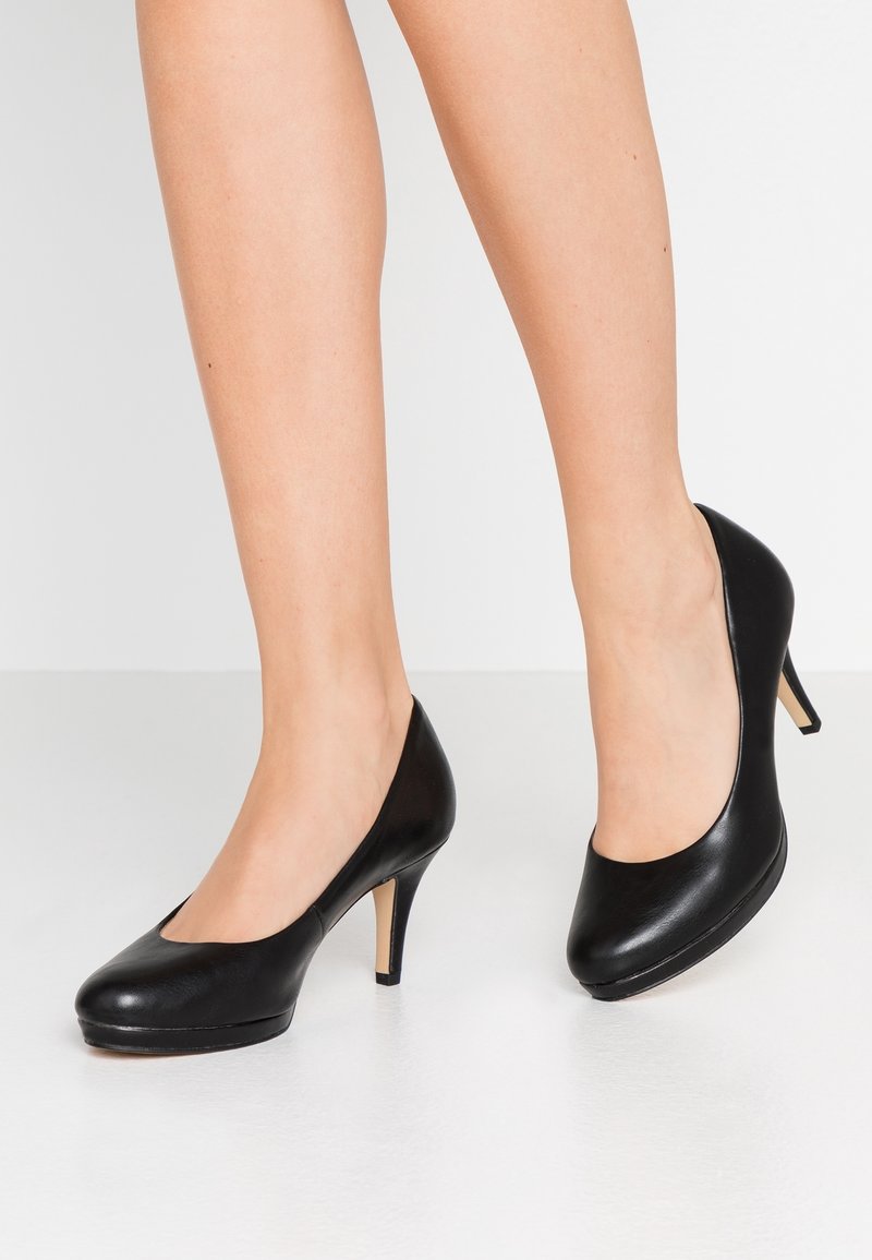 Paire de jambes portant des escarpins classiques en cuir noir à talons hauts avec un bout arrondi, posées sur un sol gris clair.