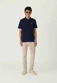 Camisa polo azul-marinho com mangas curtas, apresentando um colarinho e um pequeno logotipo. Combinada com calças bege claras e ténis claros, estilizada com óculos de sol.