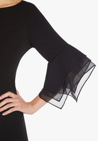 Robe noire avec des manches évasées en tissu transparent noir. Texture lisse et corsage ajusté, mettant en valeur des détails de design élégants.