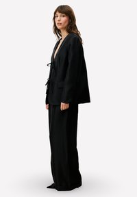 Zwart tweedelig ensemble met een diep V-hals jasje met een striksluiting en brede pijpen broek. Soepele stof en een ontspannen pasvorm.