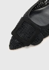 Chaussure plate noire avec un bout pointu, un délicat détail en dentelle florale et une boucle en suède noir texturé.