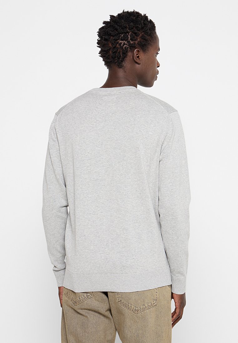 Homme avec des cheveux bouclés courts portant un pull long gris clair et un pantalon beige, tourné vers l'arrière devant un fond blanc uni.