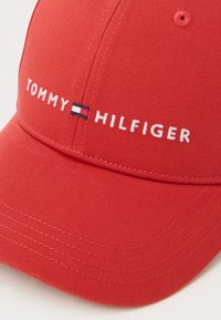 Sarkans beisbola cepures ar "Tommy Hilfiger" izšūtu baltā krāsā, un zīmola karoga logo starp vārdiem uz priekšējā paneļa.