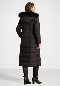 Duga crna dukserica s umjetnom krznom na kapuljači, kvadriranog dizajna, s bočnim prorezima, u kombinaciji s crnim čizmama do koljena s potkoljenicama.